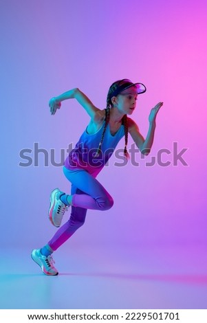 Running technique. Sportive girl, junior runner in stylish sportswear and cap posing isolated on gradient pink-blue background with neon filter, light. Concept of sport, fashion, fitness and education
