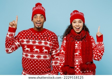 Merry young couple friends two man woman wear red Christmas sweater Santa hat posing hold index finger up great new idea isolated on plain pastel blue background. Happy New Year 2023 holiday concept