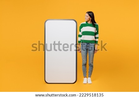 Full body smiling fun young latin woman wear casual cozy green knitted sweater look at big huge blank screen mobile cell phone smartphone with area isolated on plain yellow background studio portrait