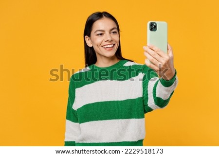 Young smiling happy brunette latin woman wear casual cozy green knitted sweater doing selfie shot on mobile cell phone post photo on social network isolated on plain yellow background studio portrait