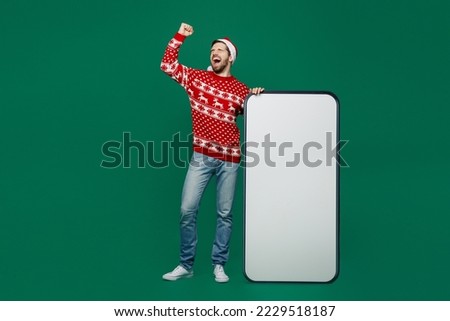 Full body merry young man wear red sweater Santa hat posing big huge blank screen mobile cell phone with area do winner gesture isolated on plain dark green background New Year 2023 holiday concept