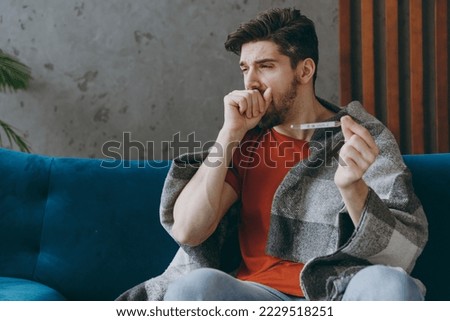 Young sick unhappy ill sad man 20s wears red t-shirt wrapped in plaid hold thermometer caugh covering mouth sit on blue sofa couch stay at home hotel flat spend time in living room indoor grey wall Royalty-Free Stock Photo #2229518251