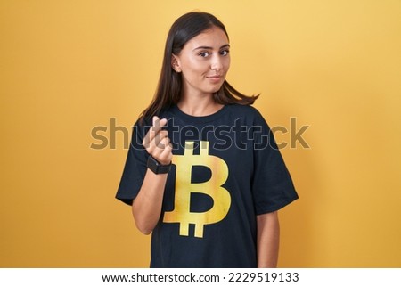 Young hispanic woman wearing bitcoin t shirt doing money gesture with hands, asking for salary payment, millionaire business 