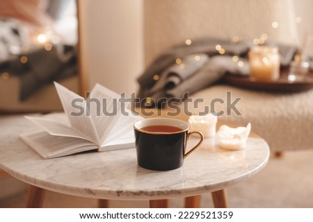 Cup of tea with paper open book and burning scented candles on marble table over cozy chair and glowing lights in bedroom closeup. Winter holiday season.  Royalty-Free Stock Photo #2229521359