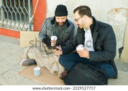 Smiling showing pictures or photos in the smartphone to a homeless man with a beard while drinking coffee together