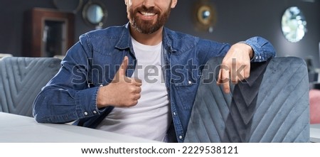 Unrecognyzable bearded man showing thumb up, while sitting at table in cafe. Crop view of smiling male in casual clothes gesturing like sign, while resting indoors. Concept of approval hand gesture.