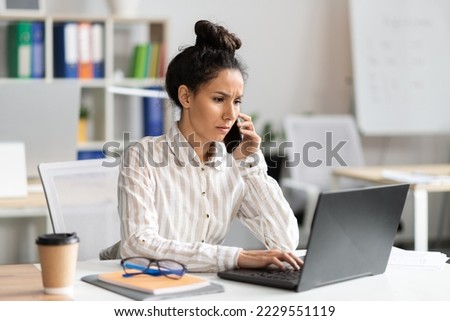 Serious businesswoman talking on smartphone and typing on laptop keyboard, sitting at workplace in office interior. Business communication and negotiation via cellphone concept Royalty-Free Stock Photo #2229551119