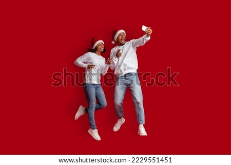 Cheerful beautiful african american couple jumping in the air, taking selfie while celebrating Christmas together, wearing Santa hats, using cell phone, isolated on red studio background, full length