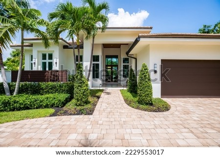 Facade of a beautiful house, with a front garden made up of palms, short grass and tropical plants, in Coral Ridge in Miami, driveway, sidewalk and street Royalty-Free Stock Photo #2229553017
