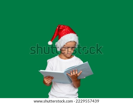 Boy in Santa Claus hat is thinking and looking at an open book, which he holds with his hands. Green background with space for text. Picture for articles and advertisements about children, holidays.