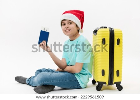 Isolated portrait of a happy preteen boy wearing Santa hat, blue t-shirt and jeans, leaning on a bright yellow suitcase, holding a passport and flight ticket, smiling a toothy smile looking at camera