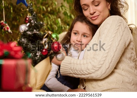 Beautiful woman, loving mother gently embracing, hugging her daughter, adorable baby girl with blurred Christmas tree on foreground. Magical atmosphere of winter holidays in family. Happy New Year