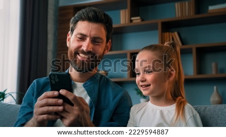 Cute kid preschool girl child looking at smartphone screen with father adult man make selfie photo together use funny face mask app on modern mobile phone gadget laughing watch online video cartoons