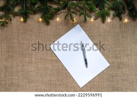 burlap background with a sheet of paper with pen and a frame of fir branches and star-shaped lights, copy space, christmas background 