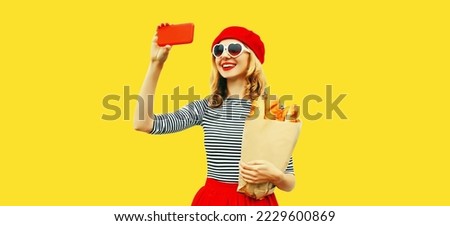 Portrait of happy smiling woman taking selfie with smartphone holding grocery shopping paper bag with long white loaf bread wearing red beret, heart shaped sunglasses isolated on yellow background