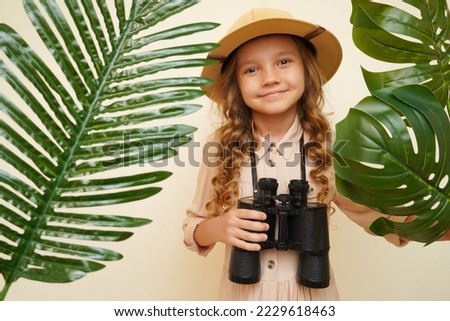 Unique child. Child blogger traveler. Kid in a safari costume. Little cute girl among palm leaves with binoculars. Child discoverer explorer traveler. Looking for treasure  Royalty-Free Stock Photo #2229618463