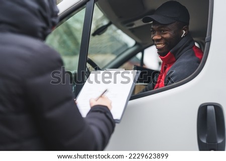 African-American delivery truck male driver smiling at unrecognizable person signing delivery papers placed on clipboard. White delivery van. Positive human interactions. High quality photo