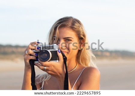 Portrait of pretty woman with old-fashioned camera. Female model with fair hair and in casual clothes holding camera on beach. Hobby, travelling concept