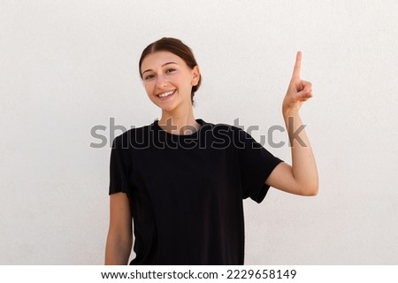 Portrait of happy young woman pointing upwards to draw attention. Caucasian woman wearing black T-shirt smiling at camera advertising something. Promotion concept Royalty-Free Stock Photo #2229658149