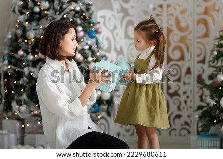 Young woman and her cute daughter girl opening a Christmas present gifts near Christmas tree indoors.