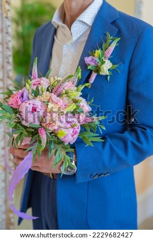 Elegant man in a suit holds a round bouquet with peonies and eustoma.Boutonniere of eremurus, white eustoma and greenery attached to grooms blue jacket. Wedding details and floristics Royalty-Free Stock Photo #2229682427