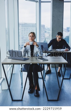 A female manager sits at a desk using a laptop in a modern office with a colleague in the background. Working atmosphere in an office with large windows.