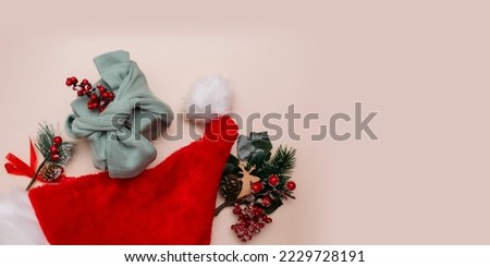A Christmas gift wrapped in olive-colored fabric and decorated with a twig with red berries lies among the fir branches and a red Santa Claus hat on a beige background. Christmas card. Copy space.