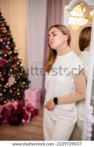 Young blond woman in a white knitted suit standing in front of a mirror in front of a Christmas tree. Winter holidays, new year