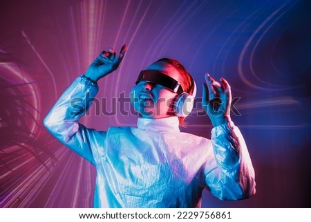 Portrait of a smiling dancing woman in futuristic sunglasses and headphones in pink and blue neon light. Music lover. Silent disco. Woman on the background of music vibes background. Futuristic Party. Royalty-Free Stock Photo #2229756861