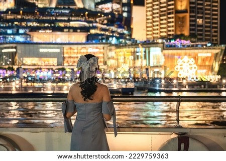 Beautiful young asian woman in dress standing and looking at illuminated department store glowing in festival event on riverside in the night. Dinner cruise sailing and sightseeing on the Chao Phraya Royalty-Free Stock Photo #2229759363