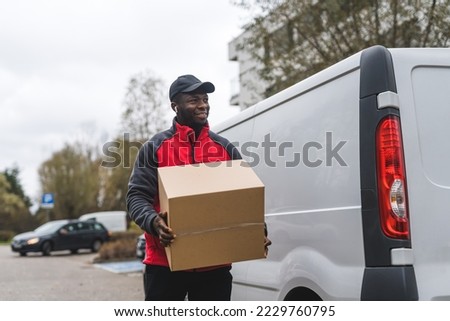 Black young adult delivery guy wearing work uniform and black cap smiling walking by white van holding carboard box package. Horizontal outdoor shot . High quality photo