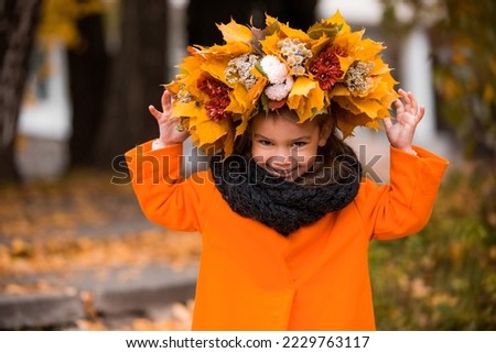 A little brunette girl in an orange coat and with a wreath on her head laughs and plays on a warm autumn day in the park