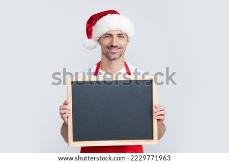 happy mature man in santa hat and apron hold blackboard with copy space