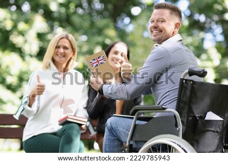 Portrait of smiling disabled man in wheelchair studying english with friends and showing thumbup sign. Happy male holding notepad with england flag. Disability and healthcare concept