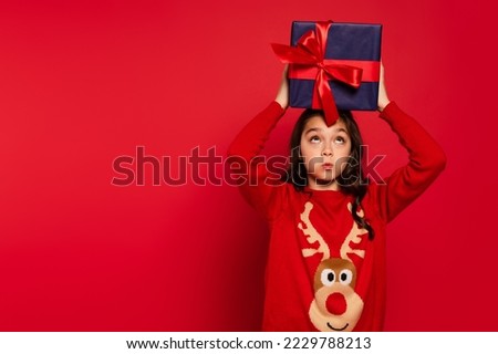 surprised child in winter sweater holding Christmas present above head on red