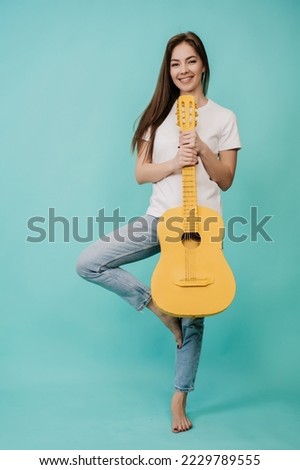 Cheerful young girl laughs, holds a yellow guitar on turquoise background in the studio balancing on one leg, dressed in white T-shirt and blue jeans, rocknroll mood. Mockup, party time, happy people.
