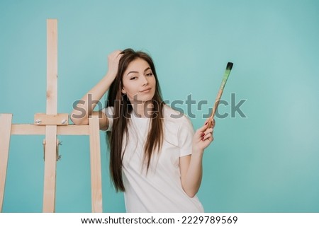 Creative blonde young woman in white t-shirt and blue jeans standing at easel with blank canvas holds paintbrush looks at camera against turquoise studio backdrop. Beautiful Italian girl paints.