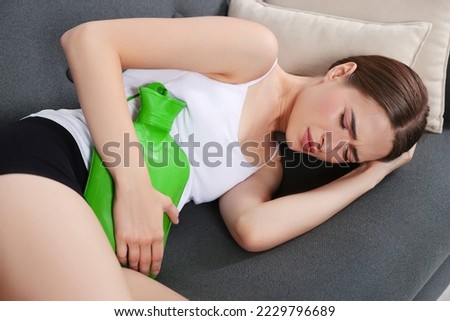 Young woman using hot water bottle to relieve cystitis pain on sofa at home Royalty-Free Stock Photo #2229796689