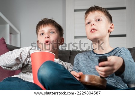 Portrait of two teenage boys sitting on sofa at home, watching interesting film movie video cartoon on TV, holding remote control, eating popcorn from red bucket and wooden bowl. Hobby, free time.
