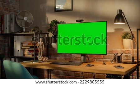 Modern Personal Computer Monitor with Green Screen Mock Up Display Standing on the Desk with Keyboard, Mouse, Pens, Notebooks and Headphones. Cozy Home Office in Loft Living Room Space.