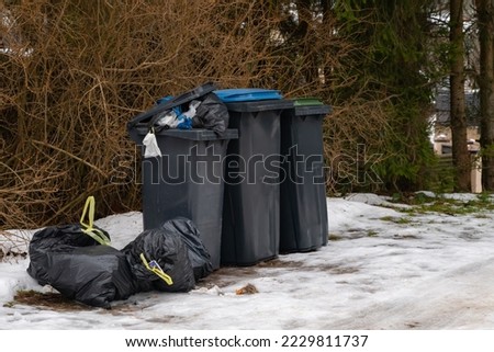 Garbage bin full of trash on white snow in residential area in winter. Royalty-Free Stock Photo #2229811737