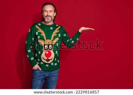 Photo of smiling confident mature man wear green ornament jumper hold palm showing new christmas brand sale season isolated on red color background