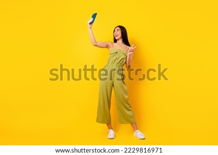 Full body portrait of cheerful adorable indonesian person hold telephone make selfie show v-sign isolated on yellow color background