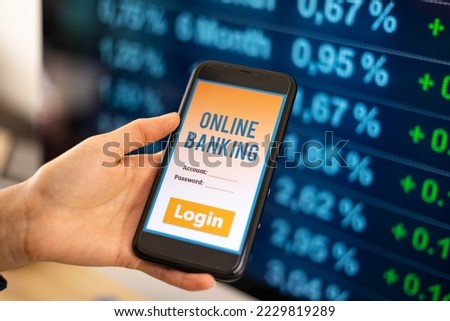 Close-up woman's hand holding a mobile phone. Online banking app with login on the screen. Monitor in the background. Business, transfer money, banking and trading concept. concept. 