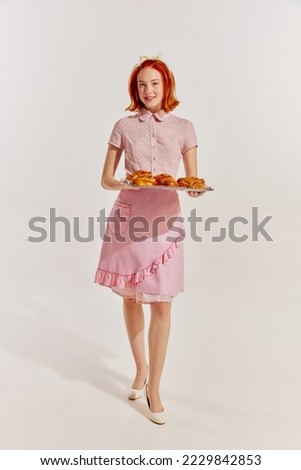 Portrait of young redhead woman in cute pink dress with freshly baked buns isolated over grey background. Concept of beauty, retro style, fashion, elegance, 60s, 70s, family. Copy space for ad Royalty-Free Stock Photo #2229842853