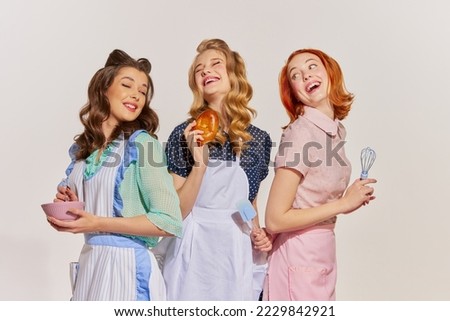 Portrait of beautiful, cheerful young women, housewives with cooking tools isolated on grey background. Pop art. Concept of beauty, retro style, fashion, elegance, 60s, 70s, family. Copy space for ad Royalty-Free Stock Photo #2229842921