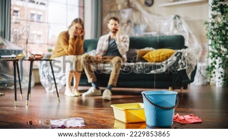 Roof is Leaking or Pipe Rupture at Home: Panicing Couple In Despair Sitting on a Sofa Watching How Water Drips into Buckets in their Living Room. Catastrophe, Distaster and Financial Ruin Royalty-Free Stock Photo #2229853865