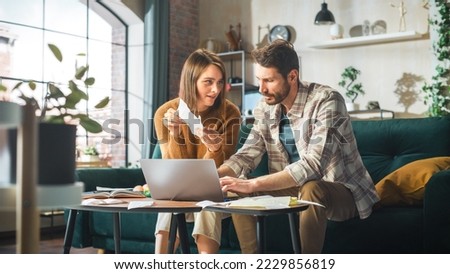 Accounting at Home: Couple Using Laptop Computer, Consulting Eachother in Apartment. Young Family Filling Tax Forms, Mortgage Documents, Bills, Checks, Balances, Invoices are in Order Royalty-Free Stock Photo #2229856819