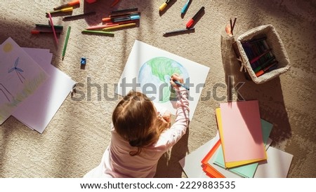 Top View: Little Girl Drawing Our Beautiful Green Planet Earth. Child Having Fun at Home on the Floor, Imagining Our Planet as a Happy Place with Clean, Sustainable Living. Cozy Sunny Day. Royalty-Free Stock Photo #2229883535