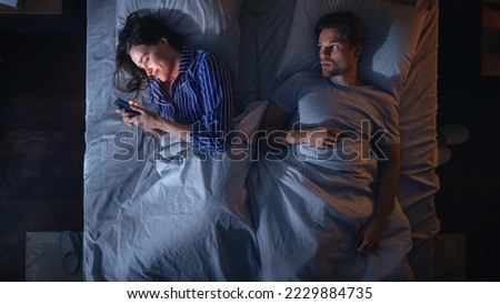 Top View Bedroom Apartment: Young Woman Uses Smartphone in Bed at Night When Her Male Partner Trying to Fall Asleep Beside. Couple Fight, Argue. Social Media, Doom Scrolling, Fake News Addiction Royalty-Free Stock Photo #2229884735